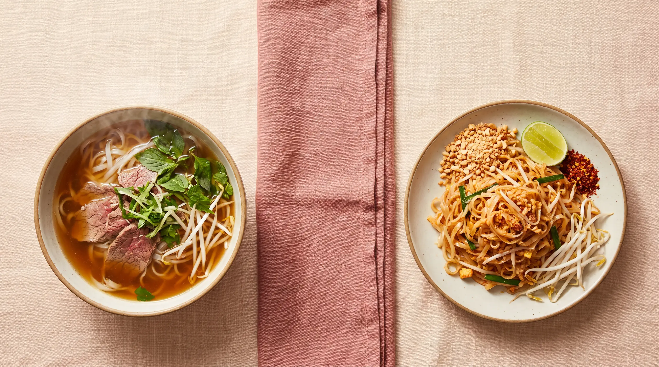 Editorial split composition: a Vietnamese pho bowl on the left with steam and herbs, a Thai pad thai plate on the right with peanuts and lime, on warm cream linen with a dusty rose napkin between them.